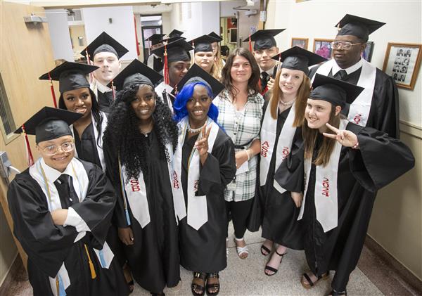 A group of graduates in their caps and gowns smile to camera with Ms. Mitchell.
